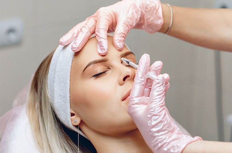 A imagem mostra uma mulher loira em uma maca, com close em seu rosto. Ela está com uma faixa branca no cabelo e com os olhos fechados. O médico está usando luvas rosas enquanto aplica Botox® entre suas sobrancelhas.